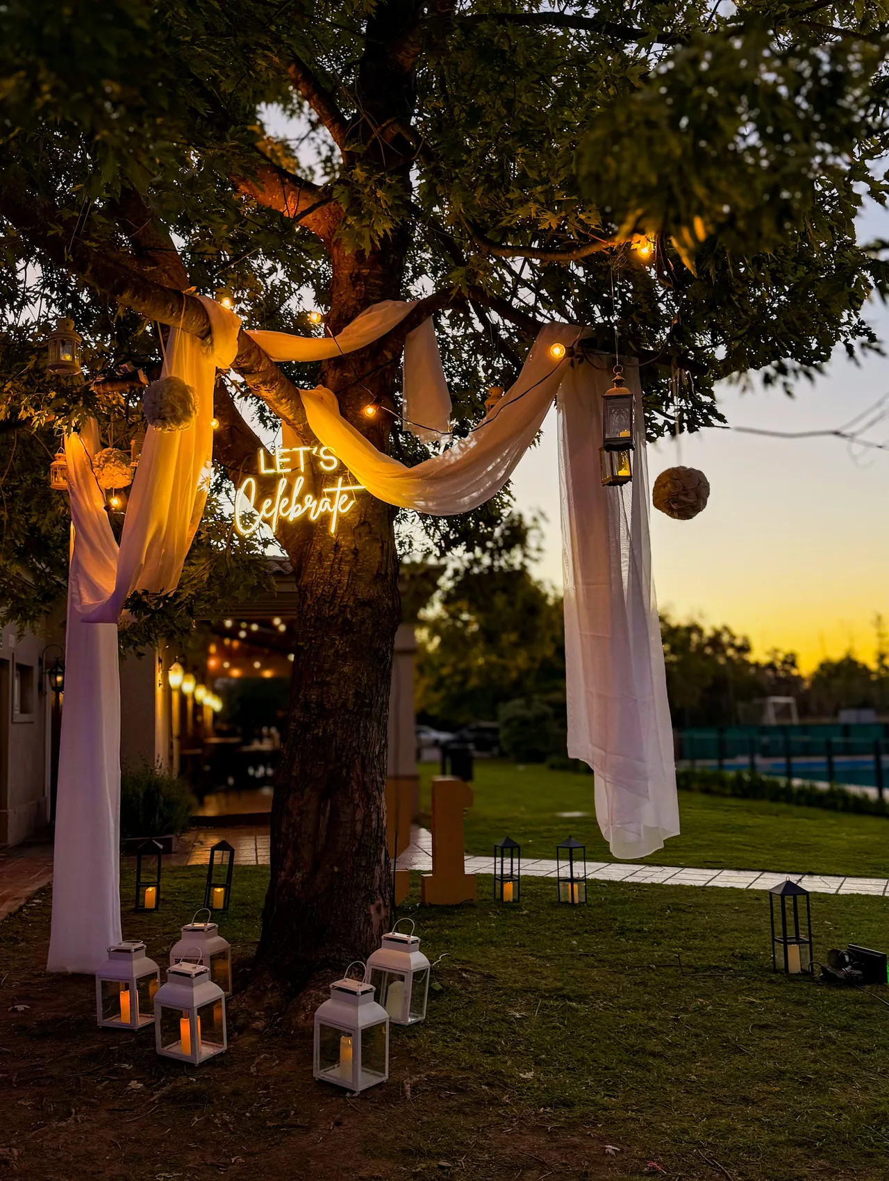 Árbol decorado con telas blancas, faroles, luces cálidas y cartel neón Let's Celebrate al atardecer