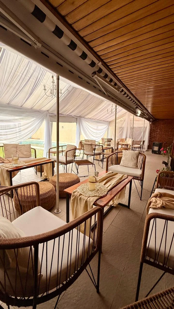 Living de ratán con pufs, mesas de madera, velas y cortinados blancos bajo carpa junto a la pileta